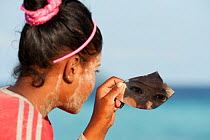 A young Bajau woman with burak (face makeup) looking at herself in a scrap of mirror. Burak is a paste made from pounded rice and tumeric and also serves as a natural sunblock. Wearing burak signifies...