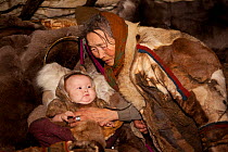 Nyaka, an elderly Nenets woman, comforts her baby grandaughter in a cradle inside their reindeer skin tent. Tambey tundra, Yamal Peninsula, Western Siberia, Russia. March 2011