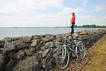 Young woman cyclist standing on stone wall viewing Rutland Water, Rutland, UK, April 2011, Model released.