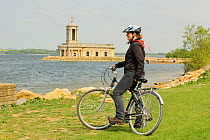 Young woman cycling round Rutland Water, stopping to view the Normanton Church Museum, a deconsecrated church that was saved from demolision when the reservior was created. Rutland Water, Rutland, UK,...