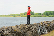 Young woman cycling around Rutland Water, standing on stone wall to admire the view, Rutland, UK, April 2011, Model released.