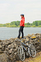 Young woman cycling around Rutland Water, standing on stone wall to admire the view, Rutland, UK, April 2011, Model released.