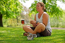 Young woman sitting on grass, hand feeding Grey squirrel (Sciurus carolinensis) in parkland, Regent's Park, London. Model released.