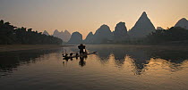 Chinese fisherman on his raft with Great Cormorant (Phalacrocorax carbo sinensis), silhouetted against the karst hills at the Li River. Yangshuo, Guangxi, China, November.