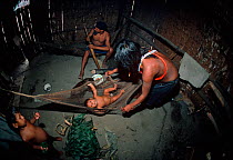 A Matses Indian family, seen inside their hut. The mother spreads out a hammock for her young baby, while another child looks on and the father works on a hunting weapon, Amazonia, Peru, November 2005...