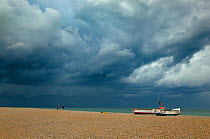 Two people walking on Cley beach near boats looking towards Blakeney Point with storm clouds overhead, Norfolk, UK, July 2011