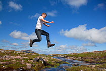 Boy (aged 17) jumping across upland stream, Cairngorms National Park, Highlands, Scotland, UK, August, Model released