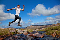 Boy (aged 17) jumping across upland stream, Cairngorms National Park, Highlands, Scotland, UK, August, Model released