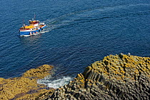 Tourist  boat approaching island of Staffa, Inner Hebrides, with columnar basalt in the foreground, Scotland, UK, July 2011