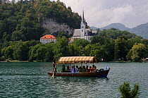 Gondolier paddling tourists in a gondola with a sun canopy on Lake Bled, passing the Succursal Church of St. Andrej, Slovenia, July 2011