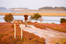 Sign for a birdwatching hide, with a birdwatcher walking towards it in the background, Arne RSPB reserve, Dorset, England, UK, September. Model released.