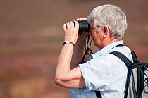 Birdwatcher looking through binoculars for visiting Osprey, Arne RSPB reserve, Dorset, England, UK, September. Model released.