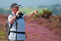 Birdwatcher looking through binoculars for visiting Osprey and pointing, Arne RSPB reserve, Dorset, England, UK, September. Model released.