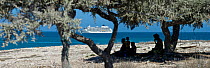 People sitting under a tree on the beach with a cruise ship in the background anchored in the Mozambique Channel off the west coast of Madagascar, near Andavadoka. Madagascar, December 2011.