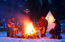 Cowboys sitting round camp fire at dusk, one playing the guitar, Wyoming, USA, February 2012, model released