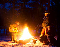 Cowboys sitting round camp fire at dusk, one playing guitar, Wyoming, USA, February 2012, model released