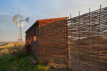 Bird hide at West Canvey Marshes RSPB reserve, with wind pump in the background, Canvey Island, Essex, England, UK, November.