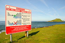 Sign warning when it is unsafe to cross a tidal causeway, with the Worm's head cut off from the mainland as an island at high tide, Rhossili, The Gower peninsula, Wales, UK, July. Sequence 2 of 2 matc...