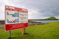 Sign explaining when it is safe to cross a tidal causeway, with the Worm's head reachable from the mainland along the wave cut platform exposed at low tide, Rhossili, The Gower peninsula, Wales, UK, J...