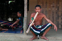 Man of Chakma tribe, smoking a bamboo pipe (daba), with woman weaving on loom in background. Tripura, India, March 2012.