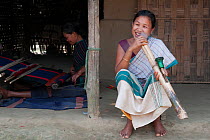 Woman of Chakma tribe, smoking a bamboo pipe (daba), with man weaving on loom in background. Tripura, India, March 2012.