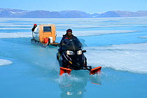 Inuit man driving snowmobile towing tourists in  sledge on sea ice, Ellesmere Island, Nunavut, Canada, June 2012.