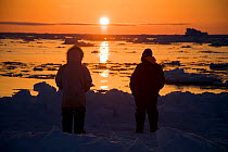Silhouette of Inupiaq subsistence whalers at the edge of an open lead in the pack ice, looking for passing bowhead whales as the mid-day sun follows the horizon. Chukchi Sea, offshore from Barrow, Arc...