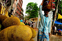 Kathal / jackfruits on a market stall, with a man walking past carrying cloths on his head, Dhaka, Bangladesh, June 2012.