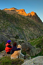 Two women hikers sitting with a Montana de los Pirineos domestic dog  at sunset watching the Remuae Valley, Parque Natural Posets-Maladeta Ribagorza, Pyrenees Huesca, Aragon, Spain. Model released.
