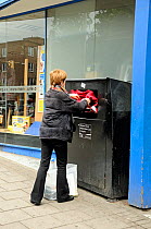 Woman recycling clothes into a clothes bank outside a charity shop, Highbury Corner, London Borough of Islington, UK