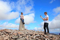 Boy taking picture of girl sitting on trig point on summit of Ben Macdui, Cairngorms National Park, Scotland, UK, August 2010.