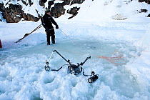 Underwater camera housing and strobes in the snow close to the 'maina' the ice diving hole, with person holding rope providing contact with the divers inside. Arctic circle Dive Center, White Sea, Kar...