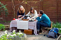 Three people sitting around a vintage sewing machine on a table outdoors. A label on the front says &#39;DIY Mending and Styling!&#39; Hackney City Farm, London, England UK, March 2011