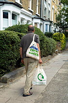 Man walking along urban street carrying two reusable cotton shopping bags with recycling logo printed on one and Think Green on the other, Holloway, London Borough of Islington, England, UK, September...