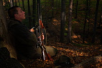 Romanian forester sitting in the forest, leaning at a Common beech (Fagus sylvatica) trunk at his shooting stand during a driving hunt for Wild boar (Sus scrofa). Mehadia, Caras Severin, Romania, Octo...