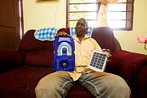 Tanzanian man at home holding wind-up radio and a portable solar panel, Miono Region, Tanzania.
