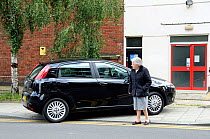 Elderly lady walks out into road as pavement is blocked by badly parked car, Holloway, London, England, UK, July 2009.