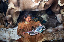 Nadia Takui, a Chukchi girl, dressed in a traditional reindeer skin Kamleika (parka) reads a magazine at the front of the polog (sleeping area) in a Yaranga (tent) at a reindeer herders' summer camp....