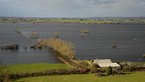 Panning shot of farm buildings at the edge of extensively flooded pasture land, with flooded farm tracks and road, West Sedgemoor, Somerset Levels, England, UK, February 2014.
