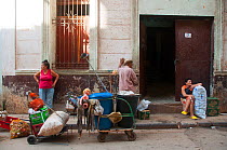 Three people on pavement including a lady leaning against wall behind bags of recycling and a wheelie bin, Havana, Cuba, October 2011.