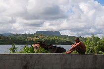 Man sitting on wall in front of Honey Bay (Baia Miel) near rusty remains of a boat with  El Yunque mountain in the distance, Baracoa, Guantanamo province, Eastern Cuba, November 2011.