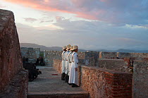Four people in traditional costumes lined up against wall, part of a Canon lighting ceremony at sunset, Fort, Santiago de Cuba, Cuba, November 2011.