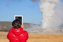 Tourist using iPad to photograph Old Faithful geyser, Yellowstone National Park, Wyoming, USA, October 2012.