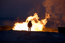 A geologist stands in front of a gas flare at dusk, at a drilling site in the South Tambey Gas Field, Yamal Peninsula, Siberia, Russia. February 2014.