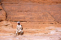 Local man sat by ancient rock engravings of human and animal figures, thought to be at least 8000 years old. Northern Niger, 2005.