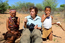 Female tourist with Naro San woman and girl eating thirst-quenching kombrua root. Kalahari, Ghanzi region, Botswana, Africa. Dry season, October 2014.