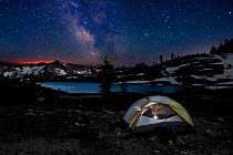 Camper inside tent reading  at night in  Snowy Lakes Basin, glow of forest fire on the horizon. North Cascades area of the Okanogan Wenatchee National Forest, Washington, USA, July 2014. Model release...