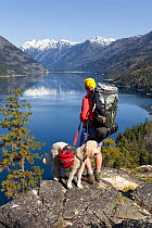 Backpacker with dog looking out over lake on the Lake Chelan Trail between Moore Point and Stehekin, Lake Chelan-Sawtooth Wilderness, Washington, USA, May 2014. Model released.