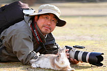 Photographer Yukihiro Fukuda, lying on the ground with a rabbit, Okunoshima 'Rabbit Island', Takehara, Hiroshima, Japan.