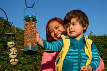 Young boy held up by his mother inspecting garden bird feeder filled with peanuts that he has just hung up, Bristol, UK, October 2014. Model released.